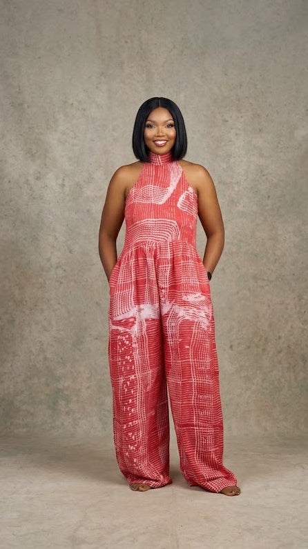A woman stands against a textured gray studio backdrop wearing a sleeveless, high-neck, wide-leg jumpsuit in a coral pink and white abstract pattern.