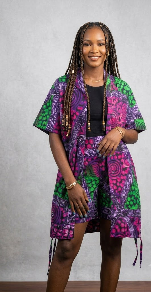 A woman modeling a short-sleeved, open-front wrap jacket with a vibrant purple, green, and pink African wax print. She is wearing a black top underneath and standing against a textured gray wall.