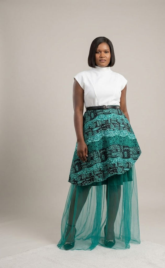 A model stands against a neutral beige studio background wearing a white sleeveless mock-neck top and a high-low skirt featuring a black and teal geometric pattern on the upper half and a flowing teal tulle lower half.